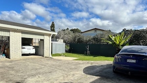 Un garage d’une maison, avec une voiture blanche garée à l’intérieur, une voiture bleue garée à l’extérieur et une clôture verte en arrière-plan.
