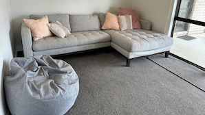 Un canapé d’angle gris avec plusieurs coussins, un fauteuil en forme de poire également gris, et une grande fenêtre donnant sur un bâtiment en pierre.