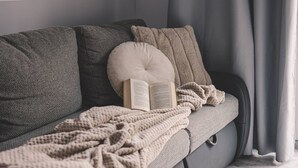 A grey sofa with a beige throw blanket and a cushion, an open book, and a window with curtains.