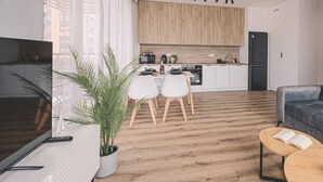 A modern kitchen with wooden cabinets, a dining area with white chairs, a black refrigerator, and a living room with a grey sofa and a coffee table.