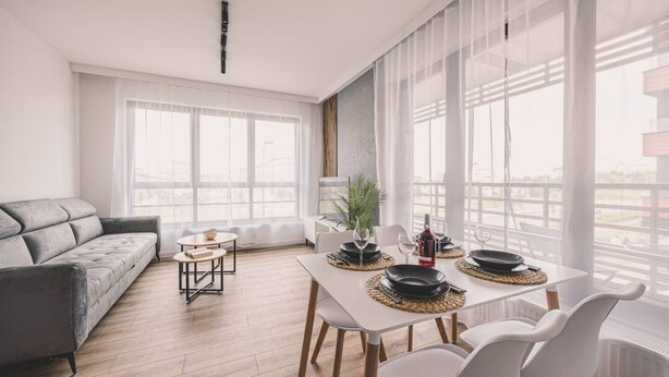 A modern living room with a grey sofa, a dining table set with plates and glasses, and large windows with sheer curtains.