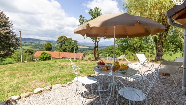 Een tafel met een beige parasol, omringd door witte metalen stoelen, op een grindpad met een landelijke omgeving op de achtergrond.