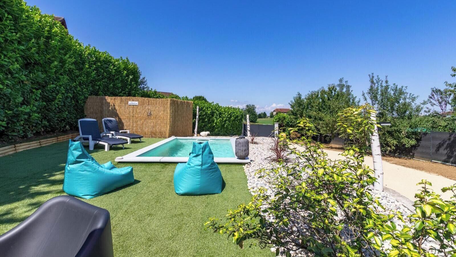 Ein Poolbereich mit Liegestühlen und Sitzsäcken, umgeben von Grünpflanzen und blauem Himmel.