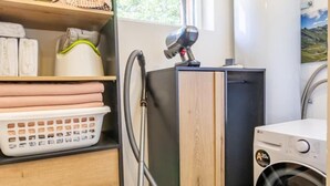 A laundry area with a washing machine, a vacuum cleaner, and a cabinet with shelves holding laundry supplies. - LE REFUGE - DOMESSIN (DOMESSIN)