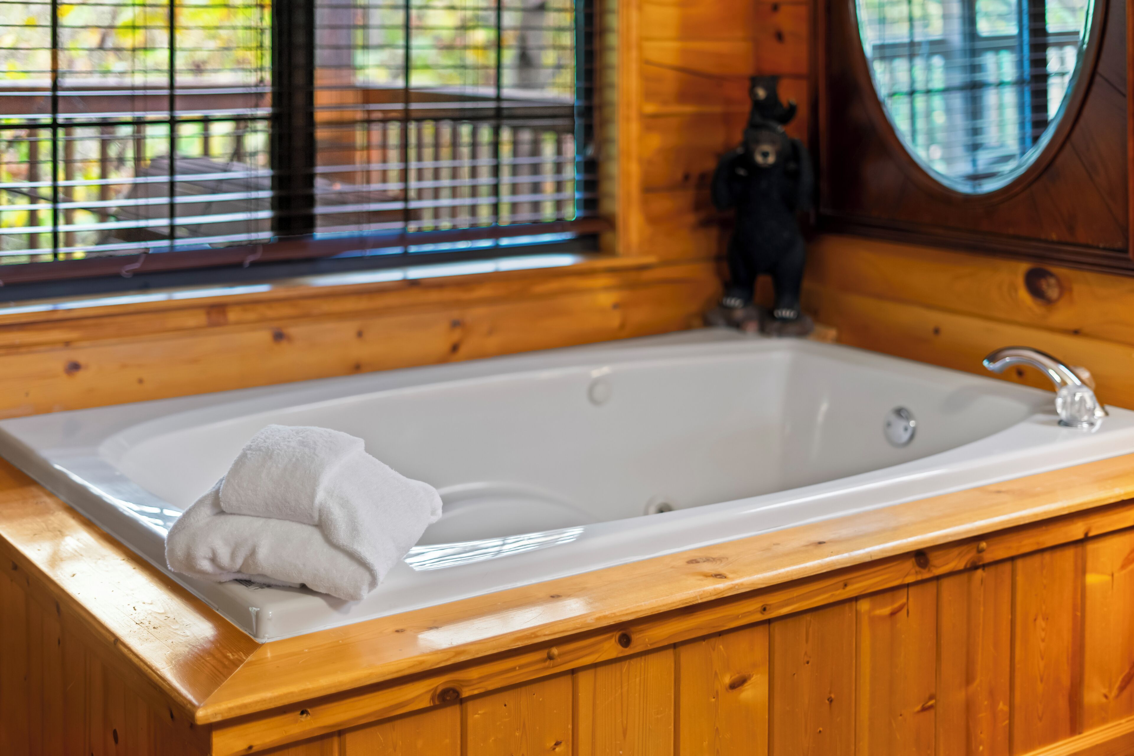 A wooden cabin interior with a large bathtub, wooden walls, and a window with blinds.