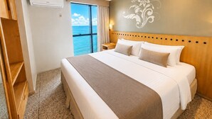 A hotel room with a large bed, a wooden headboard, a nightstand, a wall-mounted air conditioner, and a view of the sea.