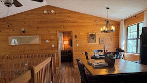A wooden interior with a dining table, chairs, a ceiling fan, and a window with curtains.