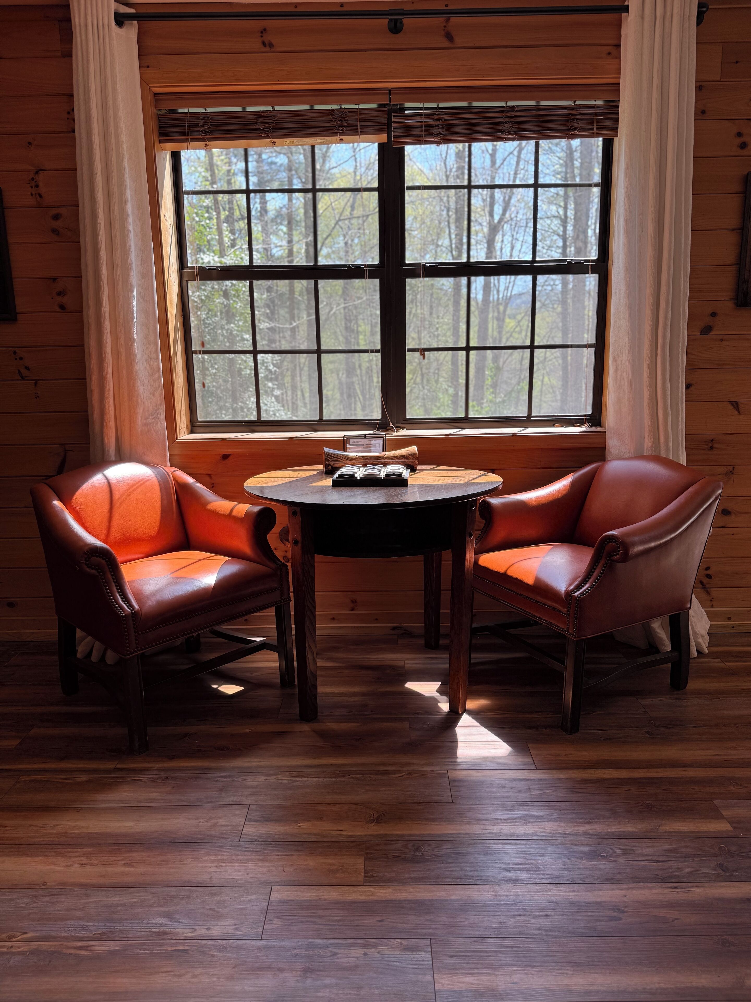 Een kamer met een houten vloer, twee leren fauteuils, een ronde tafel en een groot raam met jaloezieën.