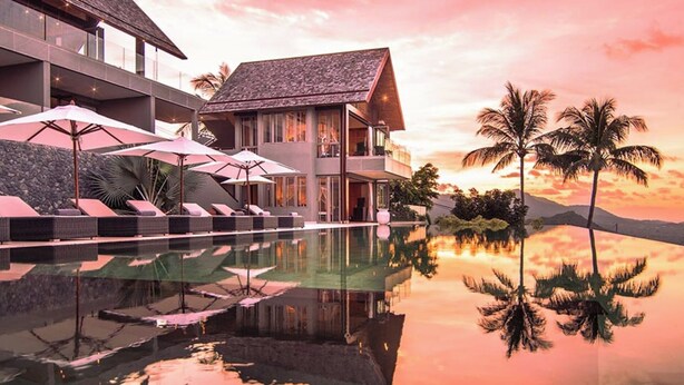 Un resort moderno con piscina que refleja palmeras y un atardecer.
