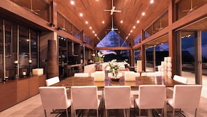 Un comedor amplio con una gran mesa de madera y sillas blancas, rodeado por paredes de vidrio que ofrecen vistas del cielo nocturno.