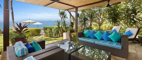 Un espace de détente extérieur aménagé avec des meubles en osier, une table à plateau de verre et une vue sur l’océan.