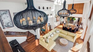 Een ruime woonkamer met een grote bank, een ronde salontafel en een eettafel met stoelen. Twee hanglampen en een trap naar een hoger gelegen verdieping.