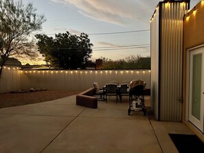Patio esterno con barbecue, sedie e un recinto bianco decorato con luci a stringa.