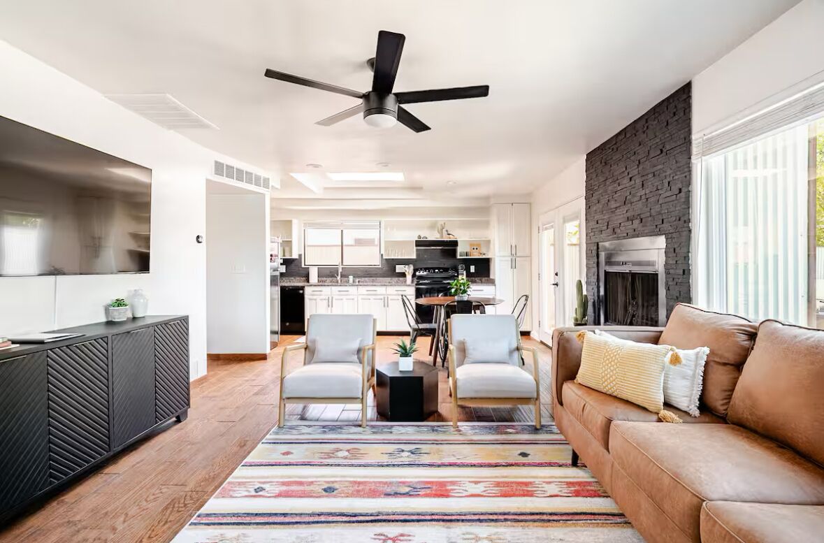 A modern kitchen with white cabinets, a black countertop, and a built-in stove. There is a dining area with a table and chairs, and a living room with a brown sofa and a ceiling fan.