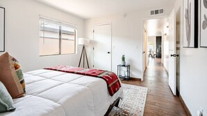 A bedroom with a large bed, a bedside lamp, a small table with a plant, and a hallway leading to another room. - Charming 3-bedroom house in Central location / UofA bike path on Mountain (Tucson)