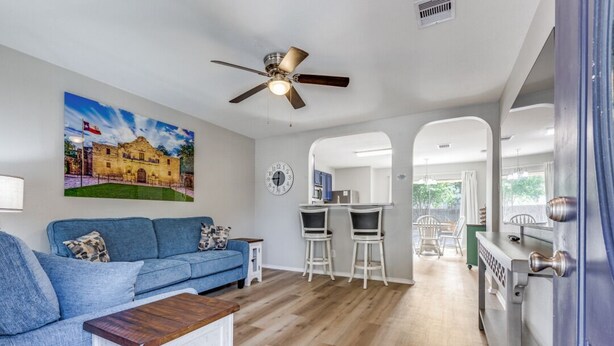 A living room with a blue sofa, a coffee table, and a painting of a building. There is a dining area with a table and chairs, and a kitchen visible through an archway. - Bright, fun, and welcoming home near Lackland and Seaworld (San Antonio)