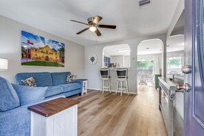Ein Wohnzimmer mit blauem Sofa, einem Couchtisch und einem Gemälde eines Gebäudes. Es gibt einen Essbereich mit Tisch und Stühlen, und durch einen Bogen ist eine Küche zu sehen.