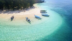 An aerial view of a beach with several boats anchored near the shore and a sandy beach with trees in the background.
