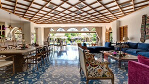 Une pièce spacieuse avec un plafond haut et un sol à motifs, comprenant un coin repas, un coin salon avec un canapé et des fauteuils, et de grandes fenêtres donnant sur l’extérieur.