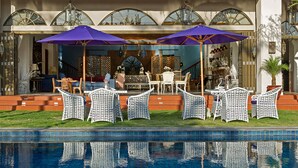 Un espace extérieur aménagé avec des chaises en osier blanc et des parasols violets, donnant sur une piscine.