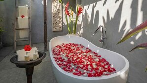 Una bañera llena de pétalos de rosa, un perchero, una mesita con artículos de aseo y un panel decorativo en la pared.