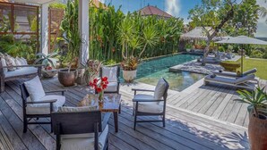 Una terraza de madera con mobiliario exterior, una piscina y abundante vegetación.