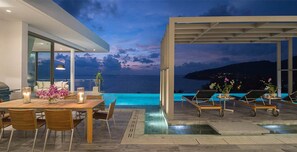 A modern outdoor dining area with a pool, overlooking the sea at dusk.