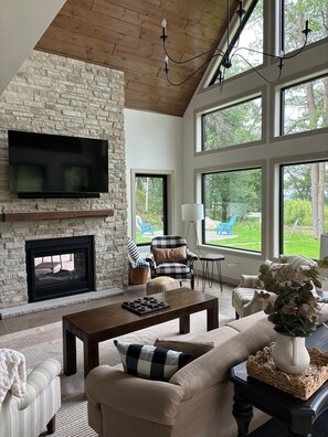 A living room with a stone fireplace, a flat-screen TV, and large windows offering a view of the outdoors. - Lake Alexander + Sleeps 4 Families + Lakeside + Fire Pit (Cushing)