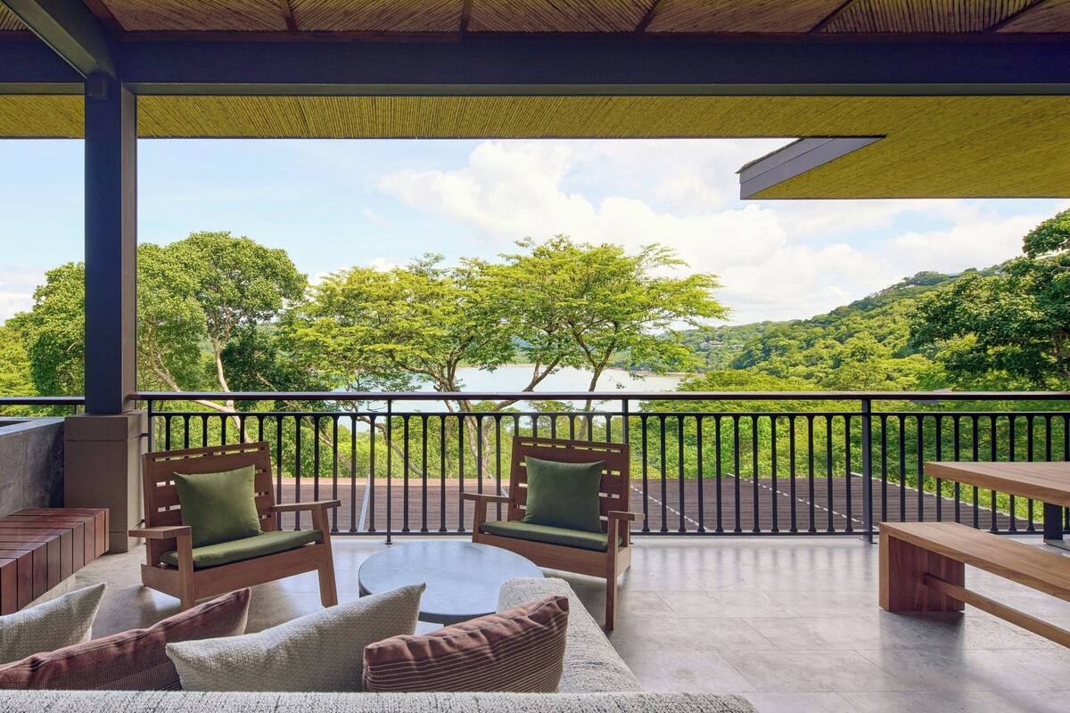 A covered outdoor seating area with wooden furniture and a view of a lush green landscape.