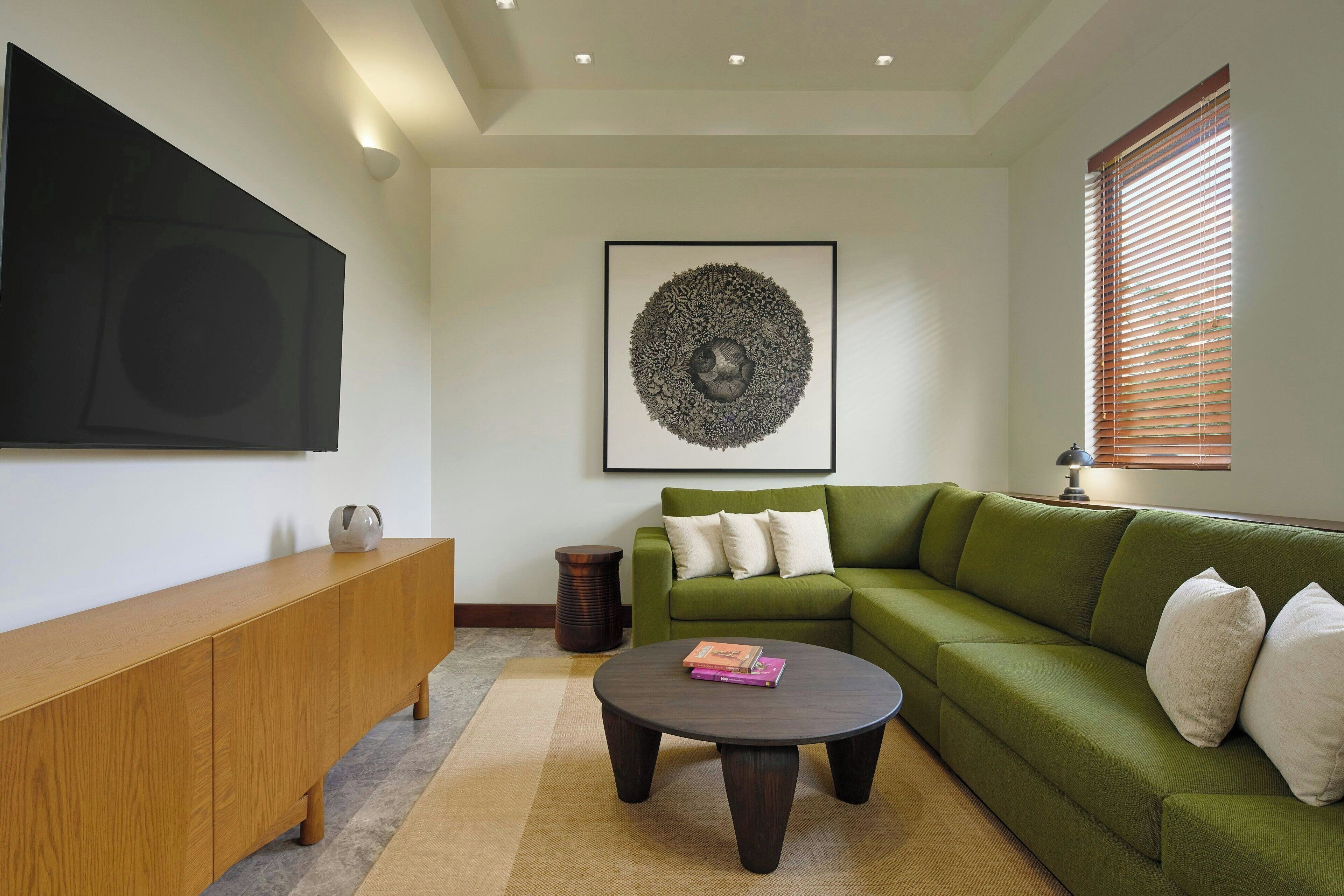 Una sala de estar con un sofá verde, una consola de madera, una mesa redonda de centro, una obra de arte enmarcada y una ventana con persianas.