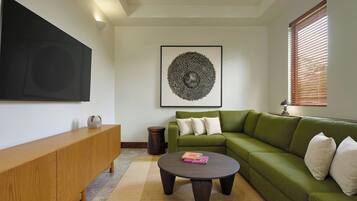 Una sala de estar con un sofá verde, una consola de madera, una mesa redonda de centro, una obra de arte enmarcada y una ventana con persianas.