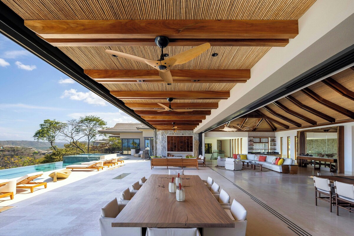 A spacious indoor area with a long wooden table, multiple seating arrangements, and a view of the outdoors.