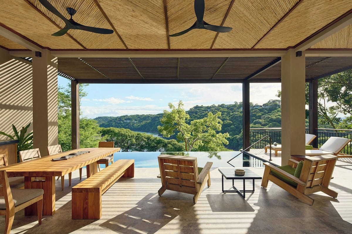 A covered outdoor area with a wooden table and chairs, a hanging chair, and a view of a lush green landscape.