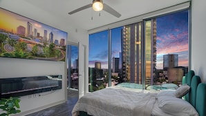 Une chambre moderne avec un grand lit, une vue sur la ville à travers de grandes fenêtres du sol au plafond, un ventilateur de plafond et un téléviseur fixé au mur.
