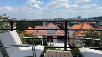 A balcony with white chairs and a table, overlooking a cityscape with red-roofed buildings and a clear sky.