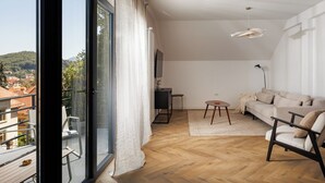 Een ruime woonkamer met een houten vloer, een bank, een kleine tafel en een televisie. Een balkon met uitzicht op de stad is zichtbaar door grote ramen.