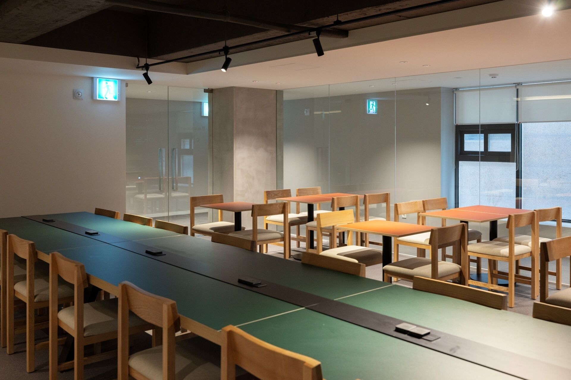 A conference room with multiple tables and chairs, glass walls, and emergency exit signs.