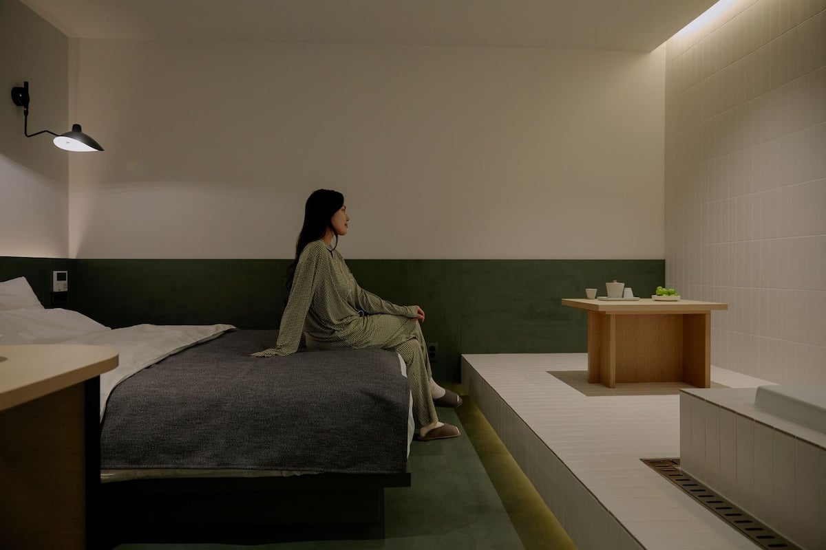 A room with a bed, a small table with cups and a plant, and a wall-mounted lamp.