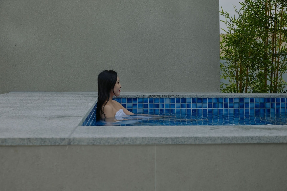 A person in a hot tub with a tiled floor and a plant beside the tub.