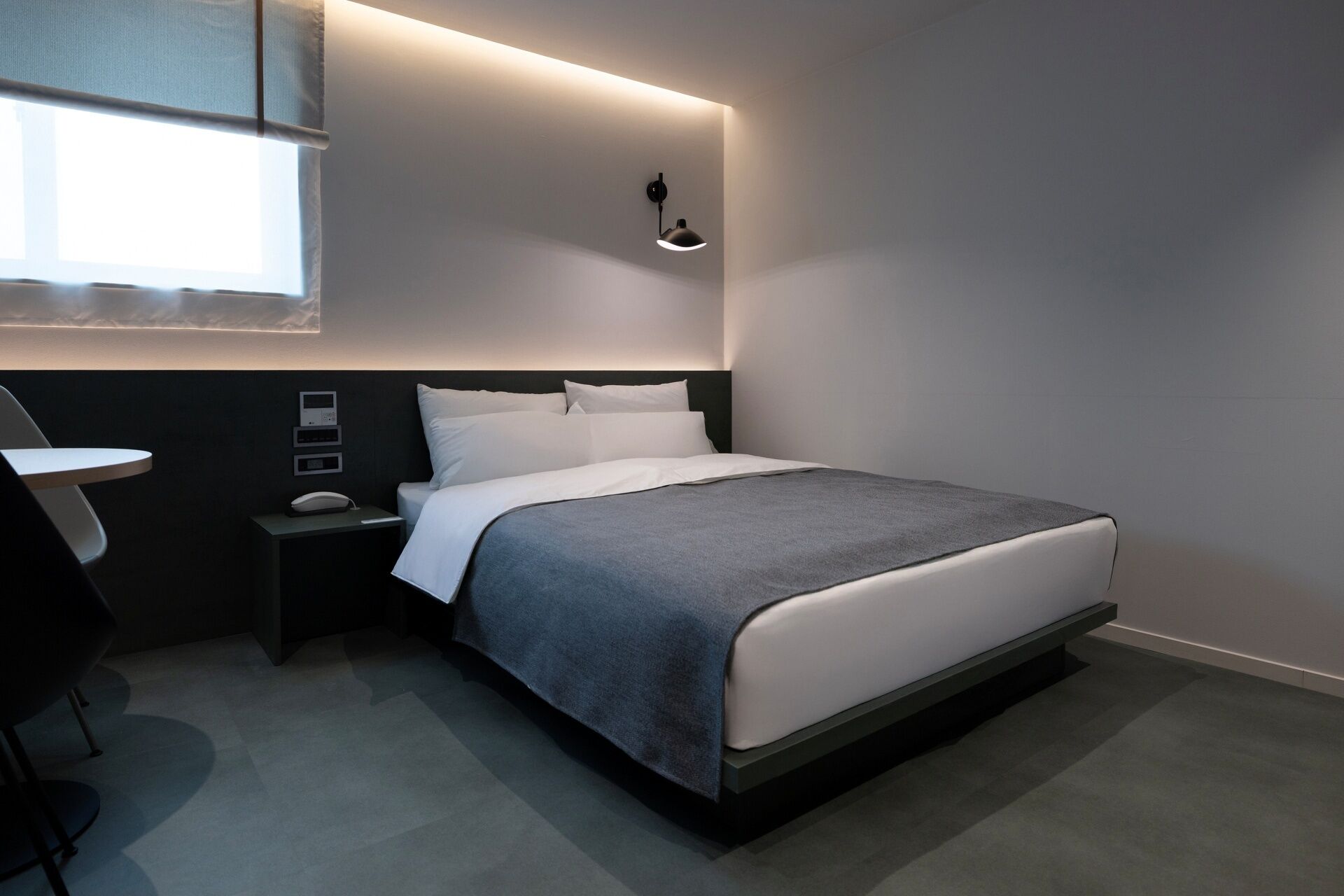 Quarto de hotel moderno com cama bem arrumada, mesa de cabeceira, abajur e janela com persianas.