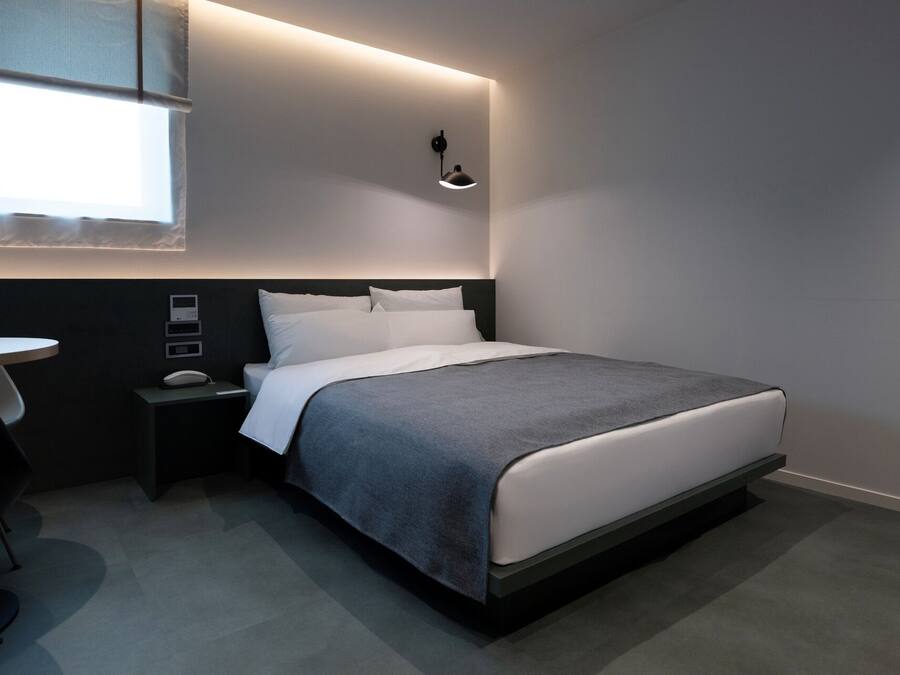 Quarto de hotel moderno com cama bem arrumada, mesa de cabeceira, abajur e janela com persianas.