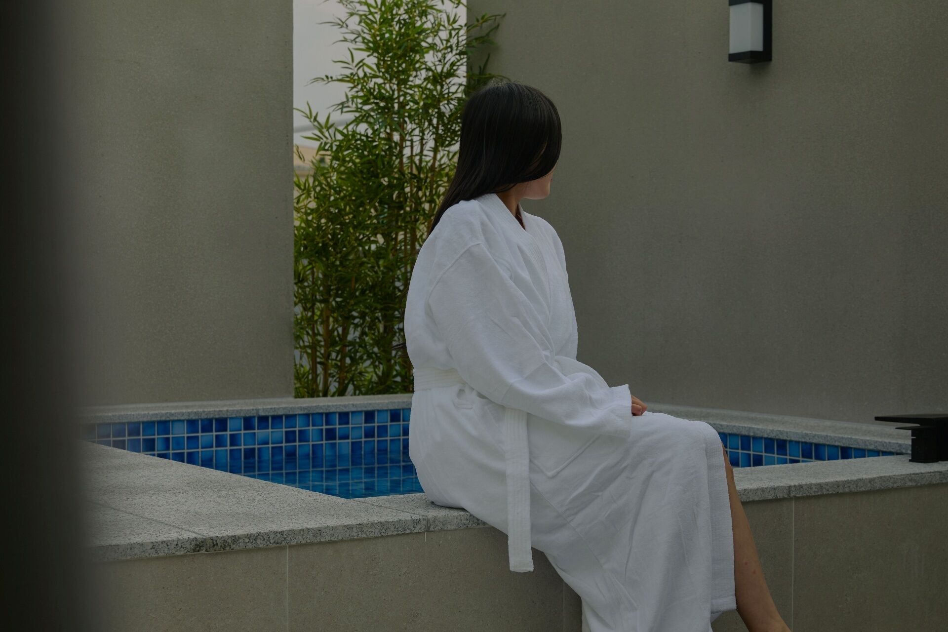 A person sitting by a pool with a tiled edge and a plant in the background.