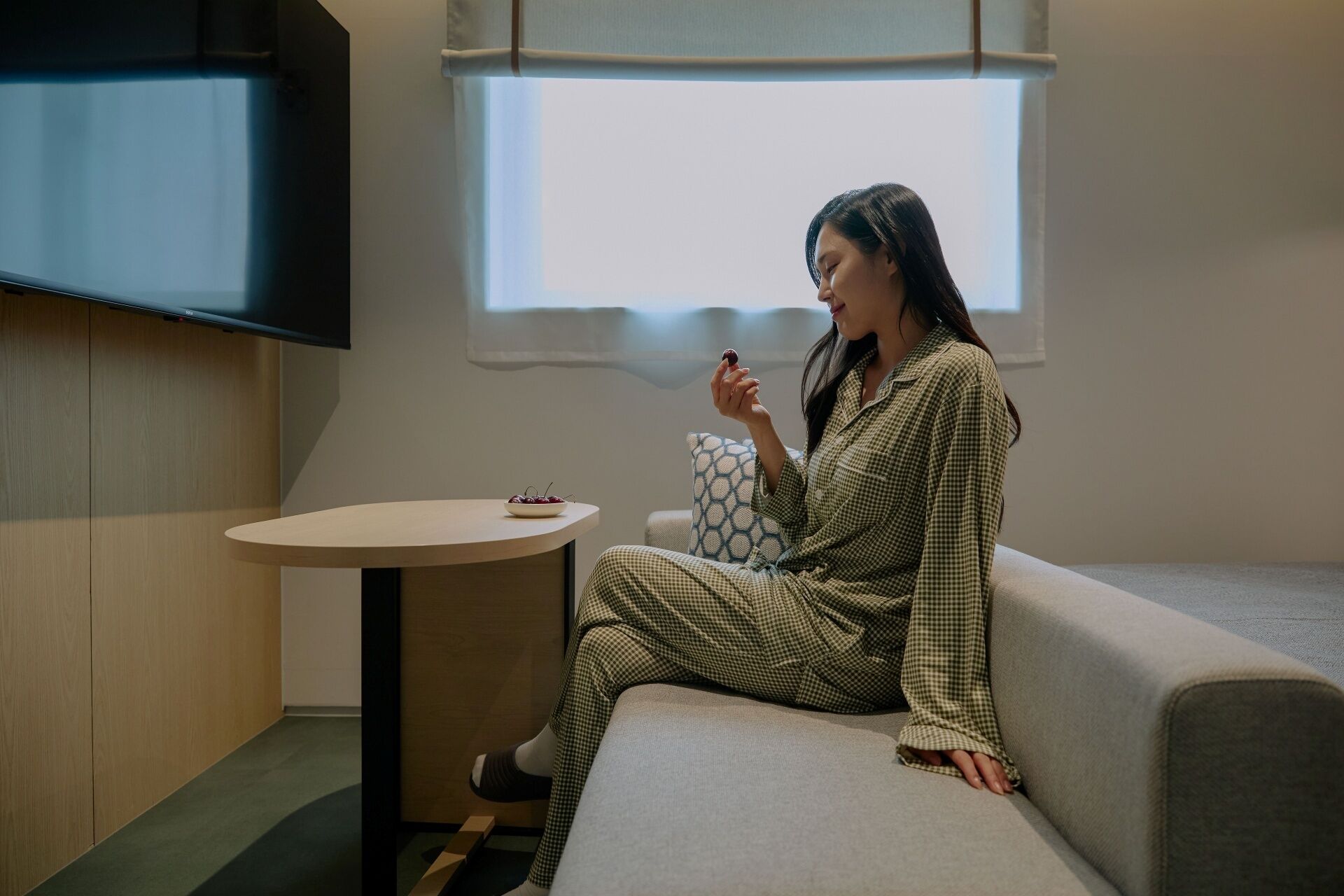 A person sitting on a sofa in a room with a television mounted on the wall, a small round table with a bowl of cherries, and a window with a view of the sky.