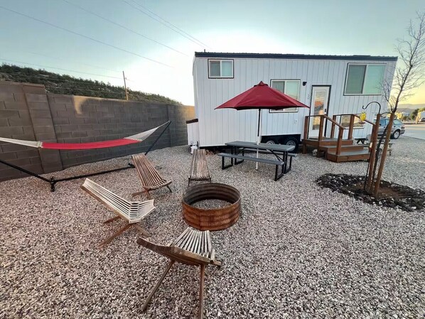 A small, single-story building with a gravel patio, a hammock, picnic tables, and a red umbrella.