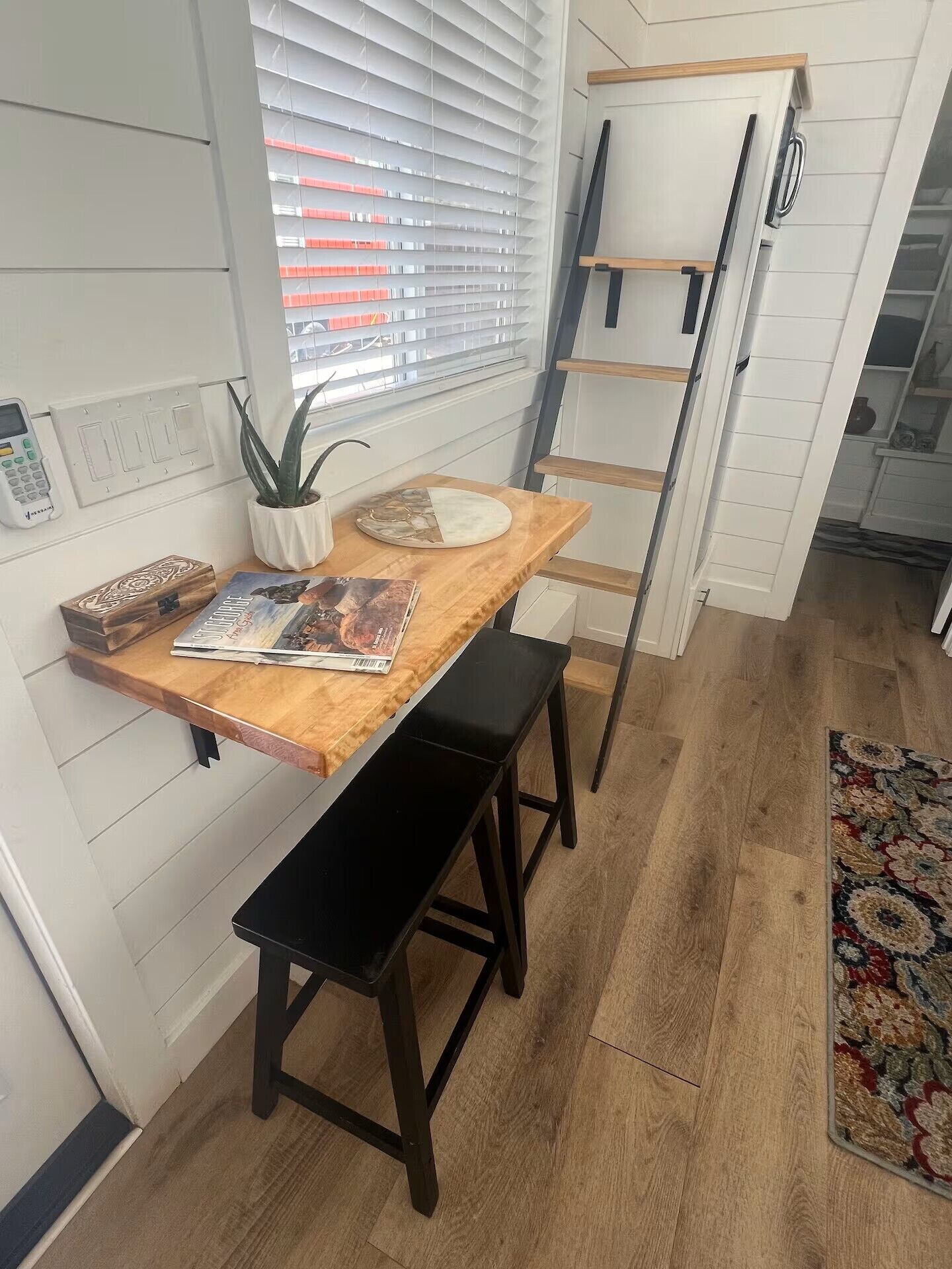 Eine Küche mit einem Holztisch, zwei schwarzen Hocker, einer Topfpflanze und einer an der Wand angelehnten Leiter.