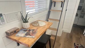 Una cocina con una mesa de madera, dos taburetes negros, una planta en maceta y una escalera apoyada en la pared.