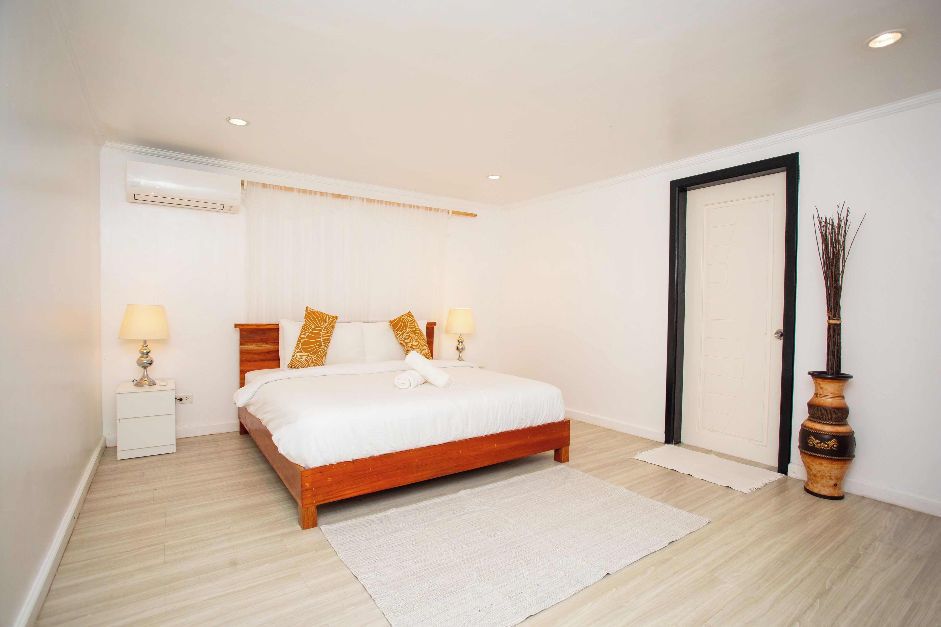 Un dormitorio con cama de madera, ropa de cama blanca, dos lámparas de noche, una mesita de noche blanca, una puerta blanca y un jarrón decorativo.