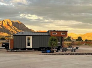 A small, modern cabin with a metal exterior and a wooden upper section, situated in a rural area with mountains in the background. - Tiny Home Lodge near Zion and Bryce Parks. Hot Tub, Stargazing, Stunning Views (Apple Valley)