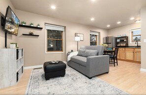 A living room with a grey sofa, a black ottoman, and a wooden coffee table. There is a kitchen area with wooden cabinets and a refrigerator. - Charming 1-bedroom Guest suite in beautiful Black Forest. (Colorado Springs)