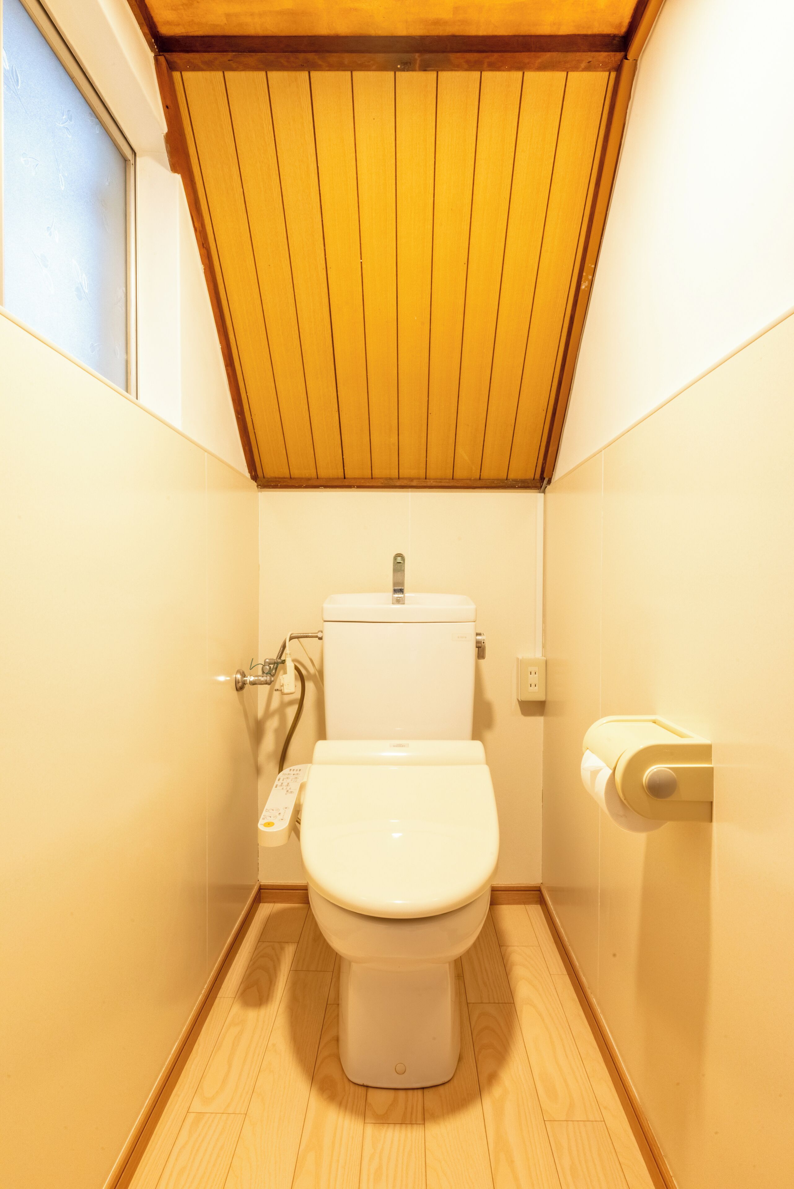 A small, compact bathroom with a wooden ceiling, a toilet, a hand dryer, and a window.